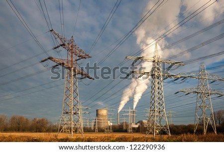 High voltage lines and power pylons in the foreground and in the background the power station with smoking chimneys and a colling tower.