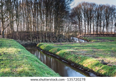 Wintry landscape with bare trees, frost on the grass and some ice on the side of the meandering ditch.