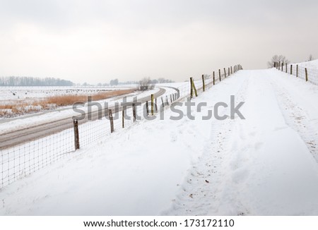 Snow-covered driveway to a dike, a country road and fences with mesh and wooden poles.