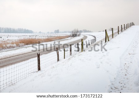 Snow-covered driveway to a dike, a country road and fences with mesh and wooden poles.