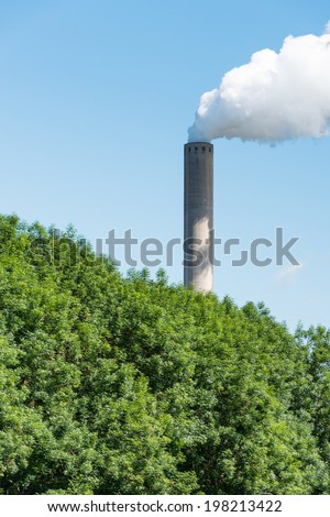 White smoke from a high chimney of a power plant hidden behind the trees.