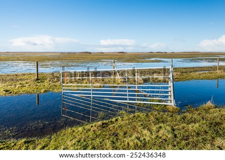 Flooded nature reserve and a crooked steel gate reflected in the mirror smooth water and ice surface on a clear and sunny day in winter.