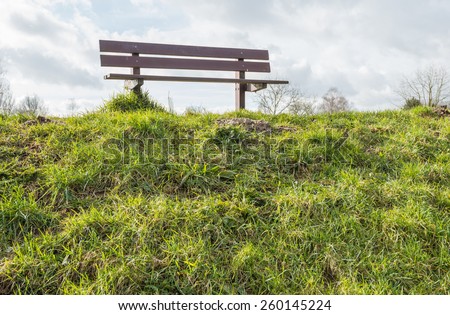 The slope of the deck is covered with grass and on top of the dike is an empty wooden bench. It is a cloudy day at the end of the winter season.