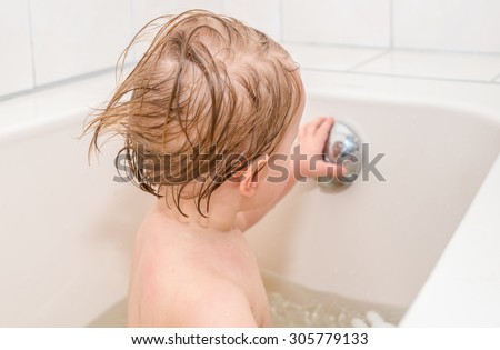 Wet baby girl with blond hair looks at her reflection in overflow drain knob of the bathtub.