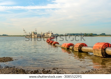 Suction dredger in a Dutch river sucks sand and gravel from the river bottom and transports it via a floating pipeline with orange floats to the shore.
