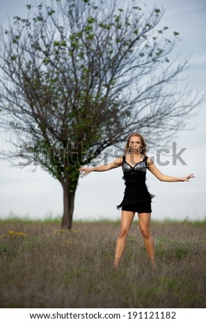 Beautiful sexy blonde girl in a black short dress posing in a field near a lone tree