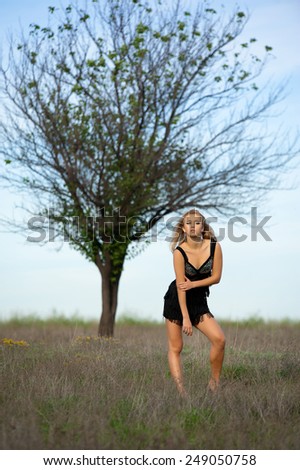 Beautiful sexy blonde girl in a black short dress posing in a field near a lone tree