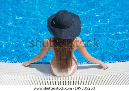 Young beautiful woman sitting on the ledge of the pool in black hat.