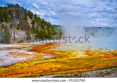 Beautiful cerulean geyser surrounded by colorful layers of bacteria, against cloudy blue sky.