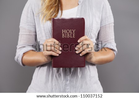 A young woman stands indoors holding a maroon bible out in front of her with both hands.
