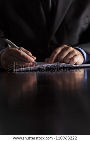 A businessman dressed in a suit sits at a desk and writes in a notebook.