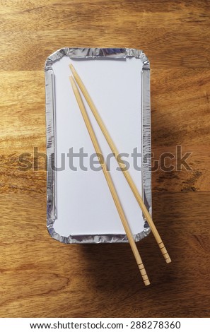 Silver foil takeaway tray with chopsticks placed on top shot from above on wooden table top