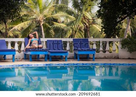 brunette girl in blue swimming suit sitting near the swimming pool with her reflection in a blue water