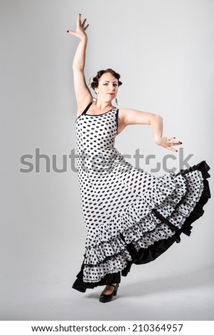 young beautiful brunette female spanish flamenco dancer in black and white flamenco dress dancing with her arms in studio on gray background