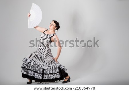 young beautiful brunette female spanish flamenco dancer in black and white flamenco dress dancing with white fan in her hands in studio on gray background