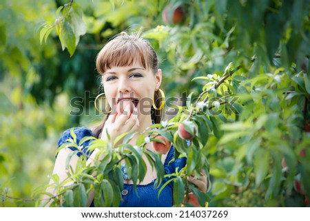 beautiful young brunette girl in blue dress eat peach in her hands stand in green garden smile and look at the camera