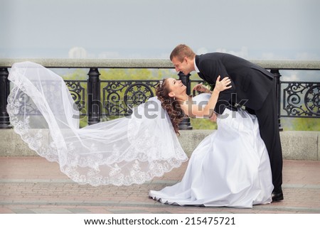 beautiful brunette bride in white wedding dress and flying veil hold her young handsome groom in black suit and stand on big city background