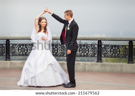beautiful brunette bride in white wedding dress and young handsome groom in black suit dance on big city background