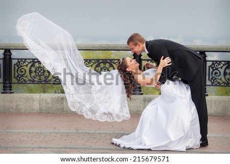 beautiful brunette bride in white wedding dress and flying veil hold her young handsome groom in black suit and stand on big city background