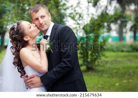 beautiful brunette bride in white wedding dress kiss her young handsome groom on green park background
