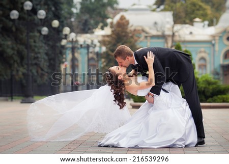 young handsome groom in black suit kiss his beautiful brunette bride in white wedding dress on big city background