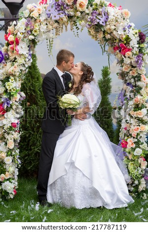young groom kiss his bride stand under floral wedding arch on marriage ceremony