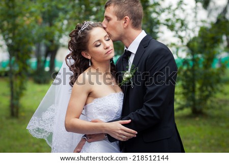 young handsome groom kiss his beautiful brunette bride in white wedding dress on green park background