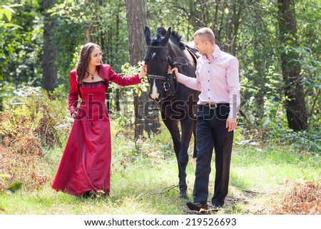 young beautiful brunette girl  in red dress walk with handsome man in pink shirt hold black horse in green forest