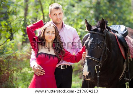 young beautiful brunette girl  in red dress hugging handsome man in pink shirt hold black horse in green forest