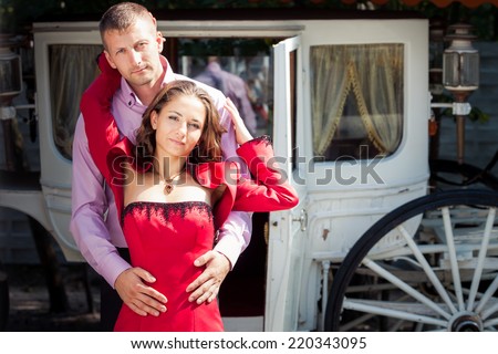 Handsome man in pink shirt hugging charming young beautiful brunette girl in long red dress and  look at the camera with white coach on background