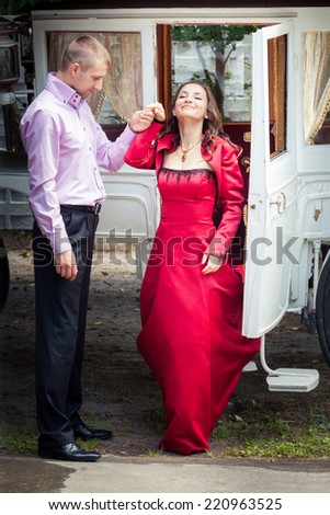 handsome man in pink shirt help young beautiful brunette girl in long red dress walk out from white coach