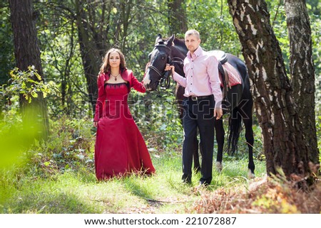 young beautiful brunette girl  walk with handsome man in pink shirt hold black horse in green forest
