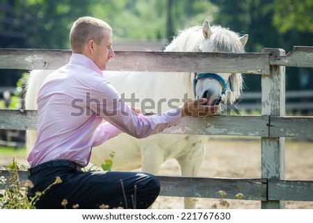 handsome man in pink shirt play with white pony horse on green field country farm