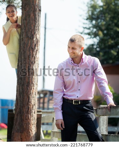 young brunette girl sit on tree and play with handsome man in pink shirt stand on green field country farm