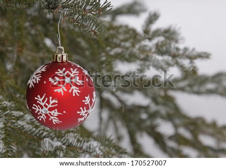 A red snow flake Christmas bauble hanging from a spruce with snow fall in the background.
