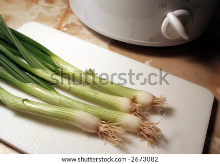 Green onions sitting on a cutting board, right next to a slow cooker.