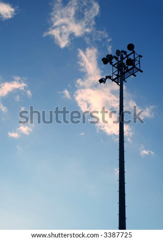 The silhouette of stadium light stand set against a blue sky with white clouds.