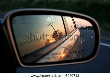 A sunset in the rearview mirror of car as a races down the road.