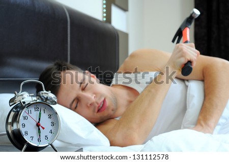 Lazy man is smashing the alarm clock with a hammer from the bed