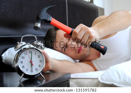 Lazy man is smashing the alarm clock with a hammer from the bed