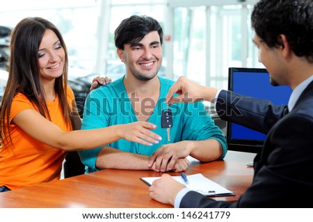 Car Dealer signing a contract to purchase and giving the key to a new car to a young attractive owners