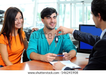 Car Dealer signing a contract to purchase and giving the key to a new car to a young attractive owners