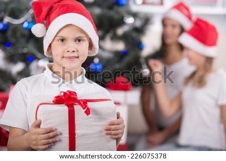 Cute boy with a gift in hands.  His family near Christmas tree in background.