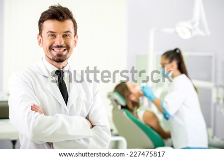 Smiling dentist is  standing in his office. In the background a woman dentist treats a patient.
