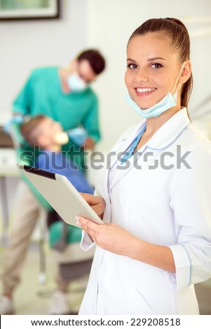 Pretty nurse with tablet, looking at camera with smile. Dentist and patient on background.