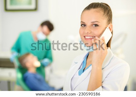 Pretty nurse is speaking by phone and looking at camera with smile. Dentist and patient on background.