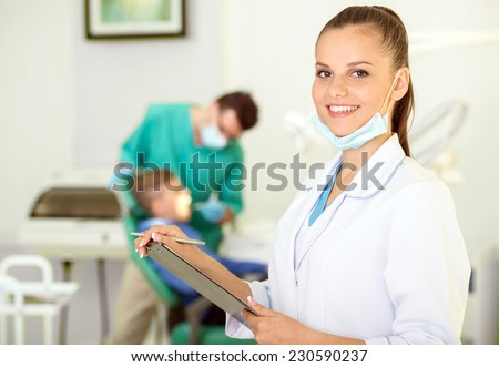 A portrait of a dental assistant is smiling at the camera with the dentist working in the background.
