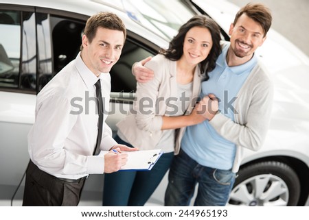 Handsome young car salesman isnstanding at the dealership telling about the features of the car to the couple.