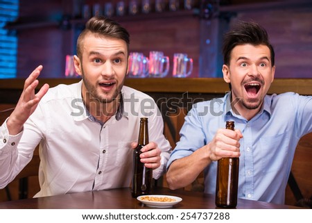 Watching TV in bar. Two happy young men are drinking beer and gesturing while sitting in bar.