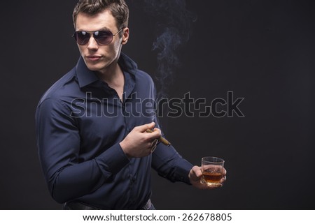 Young man in glasses is smoking a cigarette and is drinking whiskey over dark background.